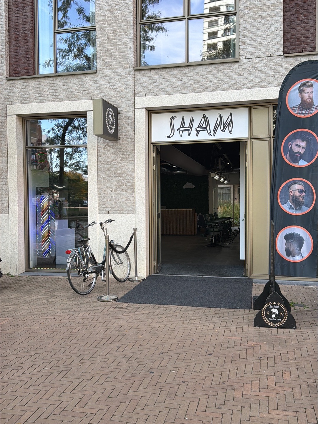 De gevel van Sham Barbershop aan de Hoflaan 3, met het SHAM-bord boven de open deur en een barberpaal in het raam